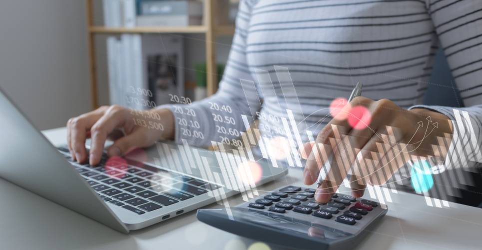 A woman doing financial work on her computer with a calculator on the desk next to her, with facts and figures displayed above it.