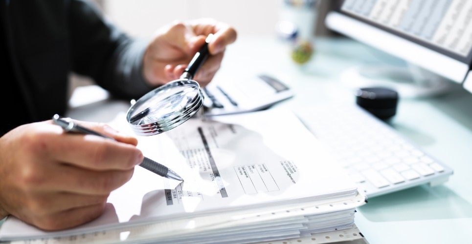 A tax expert investigating a stack of financial documents using a pen and a magnifying glass in their tax business office.