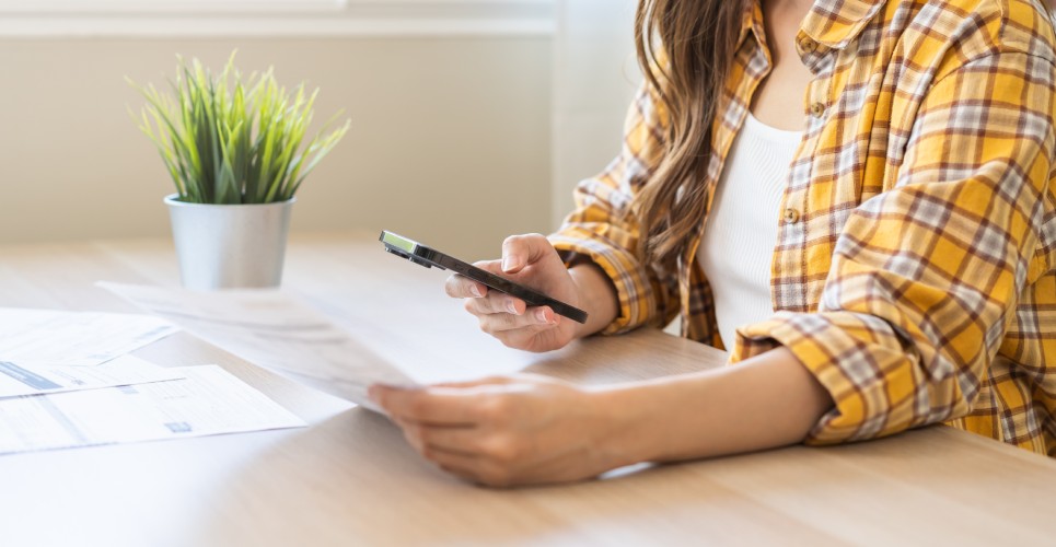 A woman in a yellow top holding her cell phone pointing toward tax documents while using a mobile tax app.