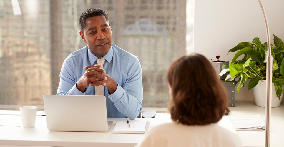 A male financial advisor sitting at a white desk and talking to a woman about her taxes with a laptop on the table.