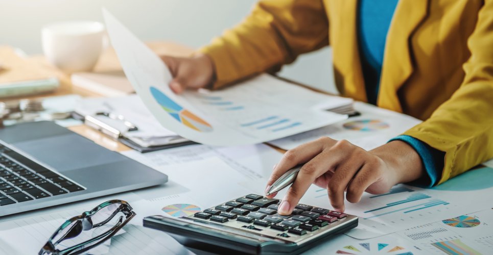 A close-up of a businesswoman analyzing a financial document in her office. There is a calculator and a laptop on her desk.