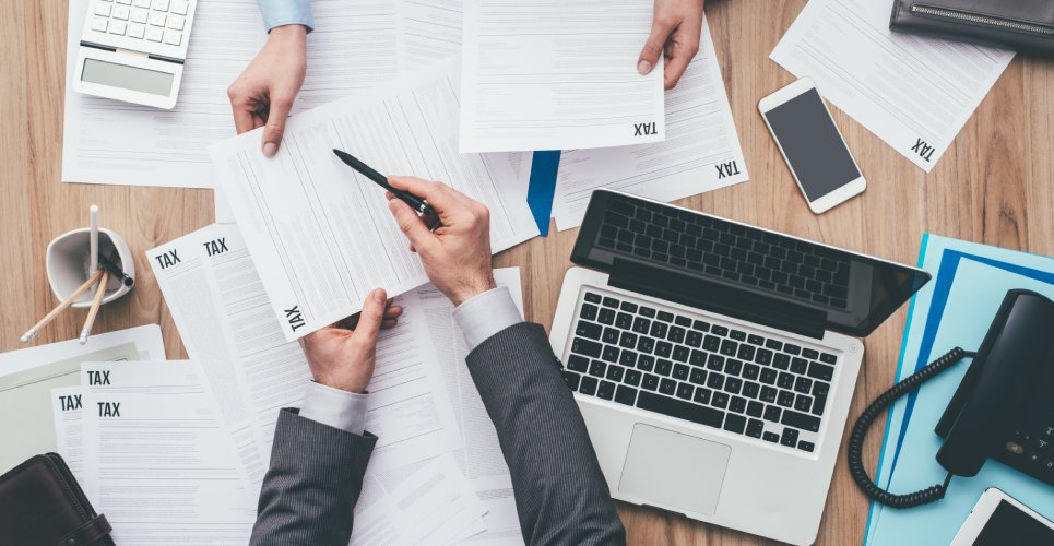 Two tax professionals handing documents back and forth over a desk. There is a calculator, laptop, and phone on the table.