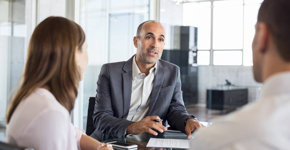 A financial advisor in a gray suit talking with two clients about their tax documents. They are sitting around a glass table.