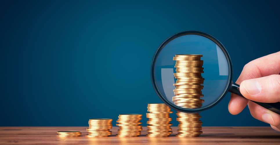 Multiple stacks of coins, increasing from left to right, on a wood table with someone holding a magnifying glass in front.