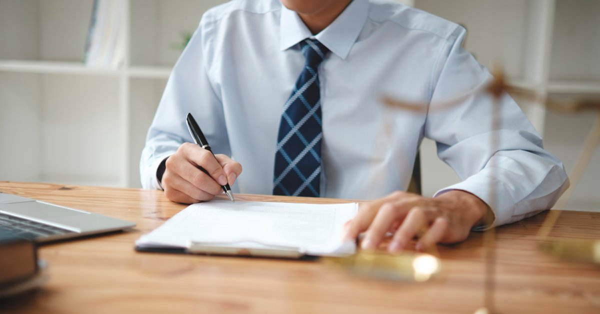 A tax service bureau employee writing with a pen on a clipboard with a laptop next to him on his desk.