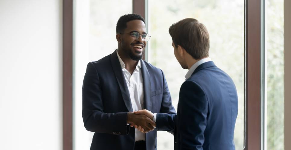 Two business professionals in dark suits smiling while shaking hands in front of floor-to-ceiling windows.