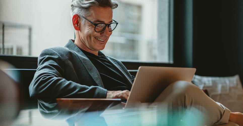 A business professional working on his laptop in an office. There is a window behind him and he is smiling at the laptop screen.