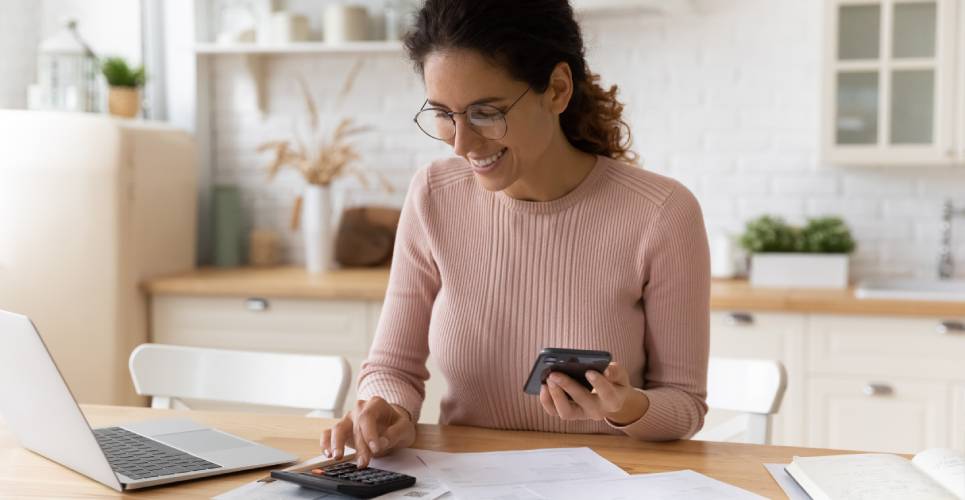 A young woman looking at financial documents while using a calculator and holding her phone. There is a laptop nearby.