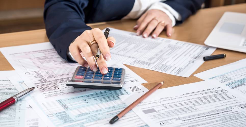 A close-up of a woman using a calculator to do her taxes. There are documents and writing utensils strewn about on the table.