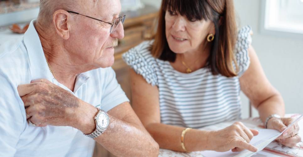 A senior couple sitting at home looking over paper documents at their kitchen table. The table has a red and white tablecloth.