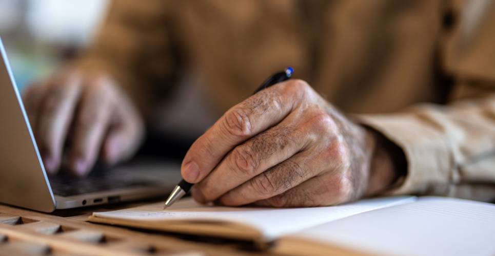 A close-up of a senior man writing something down with a pen in a journal while working on a laptop computer.