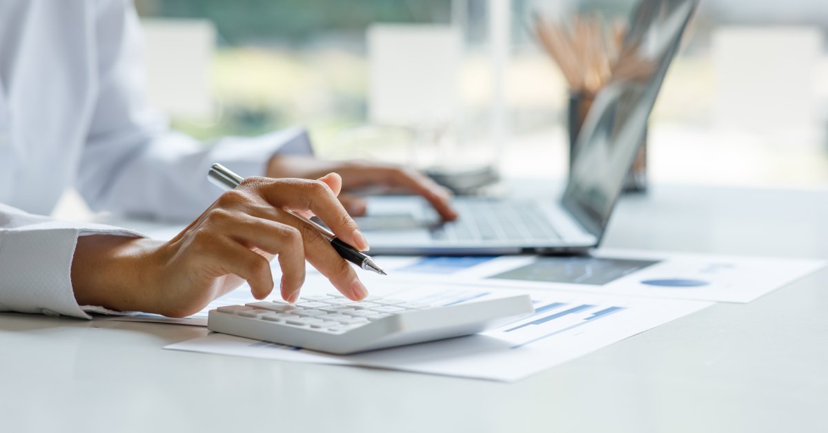 A close-up of a business professional using a calculator and a laptop while reviewing printed financial information.