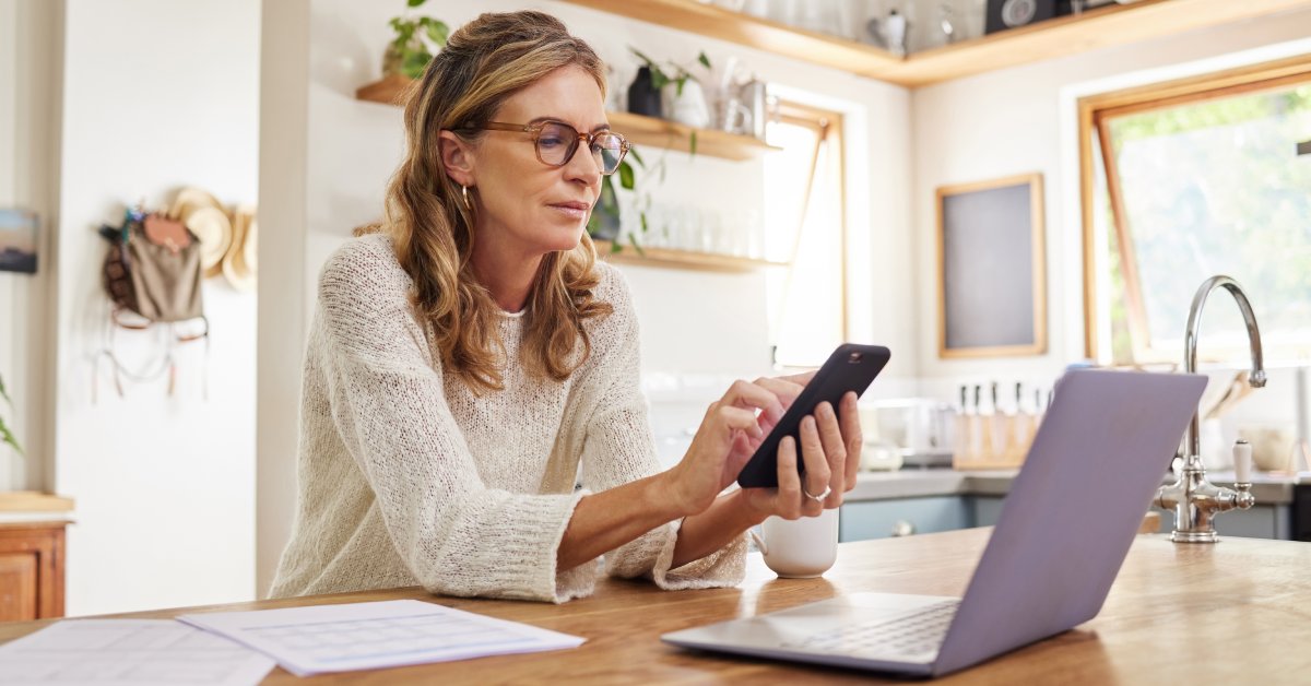 A woman with glasses looking at her phone. There are a few papers and a laptop on the table in front of her.