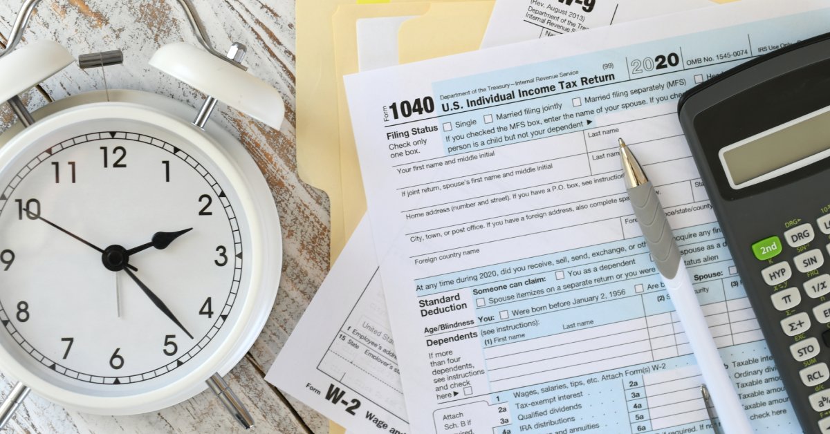 A clock on a wooden table next to a 1040 individual income tax form, ball point pen, and a black calculator.