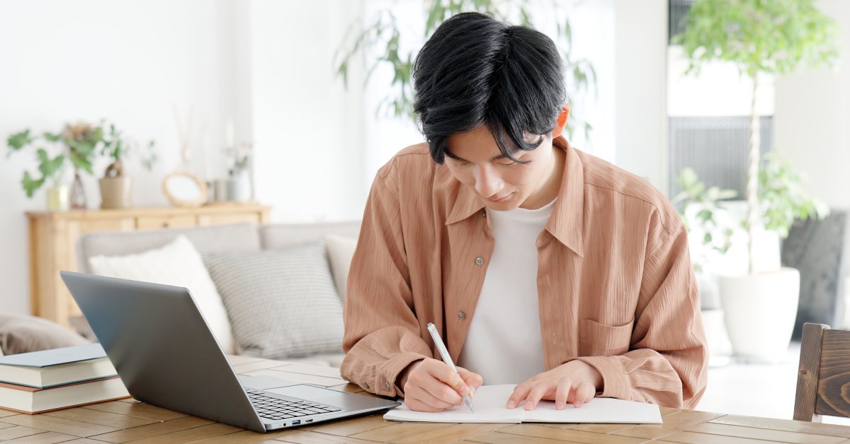 A man wearing a button down shirt and a white tee writing in a notebook while working on a laptop computer.