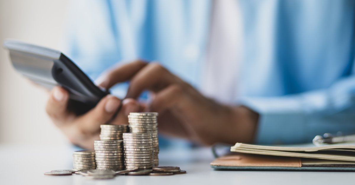 A person holding a calculator and typing numbers into it. They are sitting at a desk with a notebook and multiple stacks of coins.