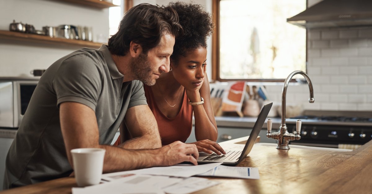 A young couple sitting together at a kitchen table and looking at a laptop. There are various papers on the table next to them.