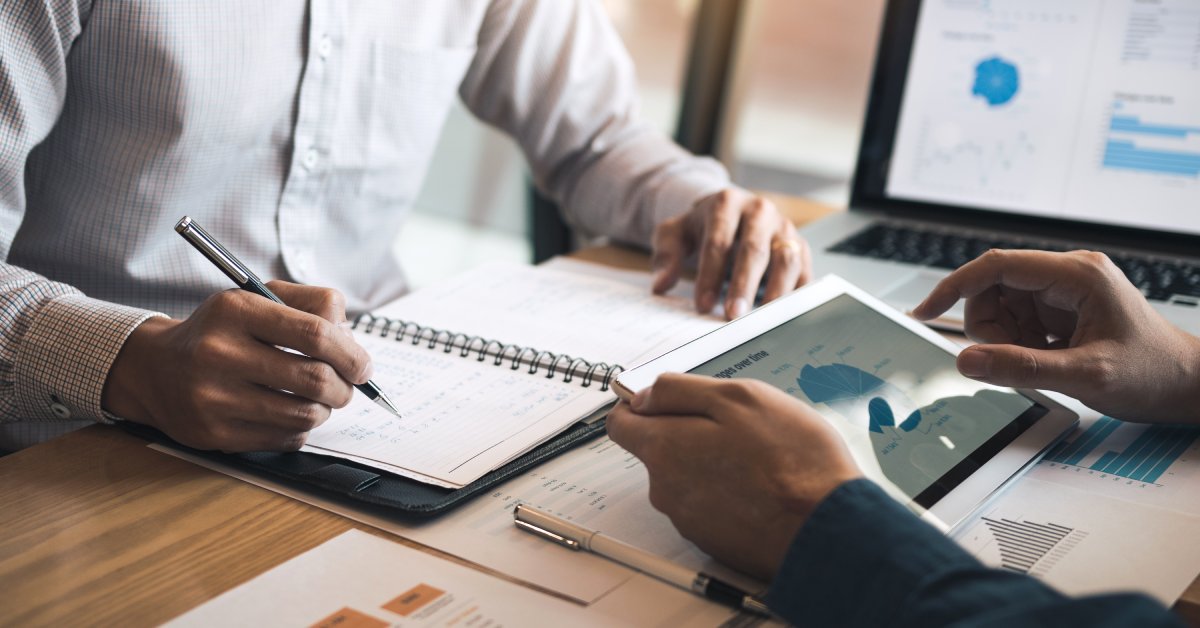 A close-up of two business partners looking at various papers and electronic devices containing financial planning graphs.