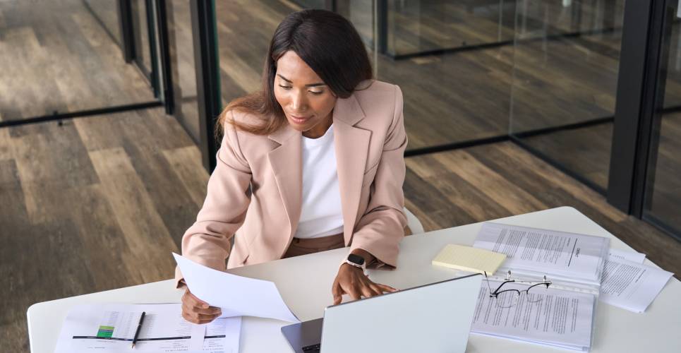 A woman sitting at a desk and looking at paper documents while working on a computer in an office building.