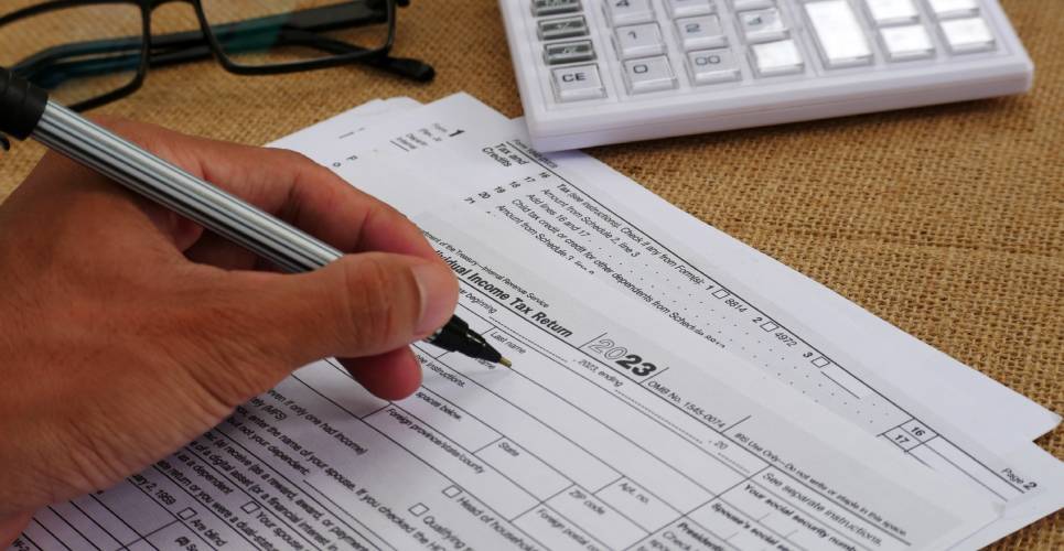 A hand holding a pen and writing on an individual tax return. There's also a pair of glasses and a calculator on the table.